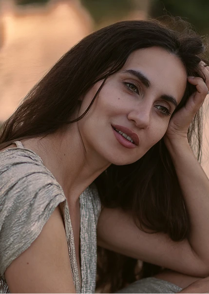 Woman with long dark hair smiling at camera in casual gray top, hand near face in relaxed outdoor portrait