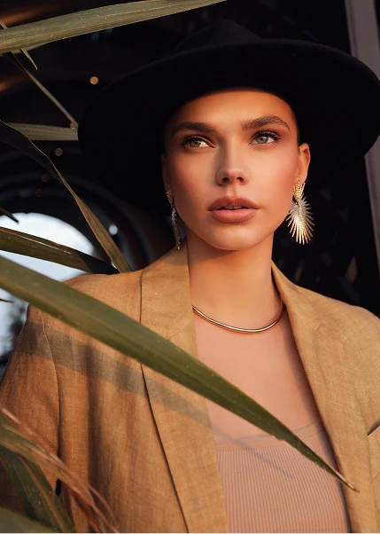 Woman wearing black fedora hat and beige blazer with gold hoop earrings and chain necklace among tropical leaves