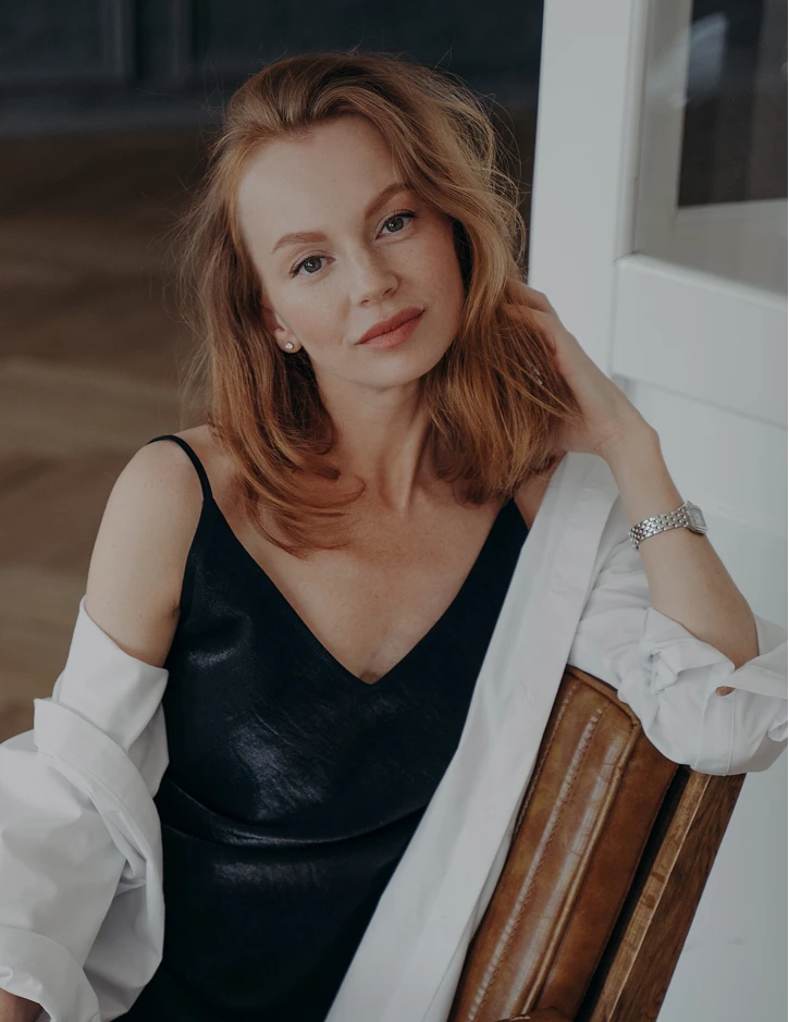 Woman in black dress with white shirt posing elegantly on wooden chair in natural lighting