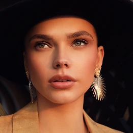 Woman in tan blazer wearing gold fan-shaped earrings with elegant makeup and dark hair against black background