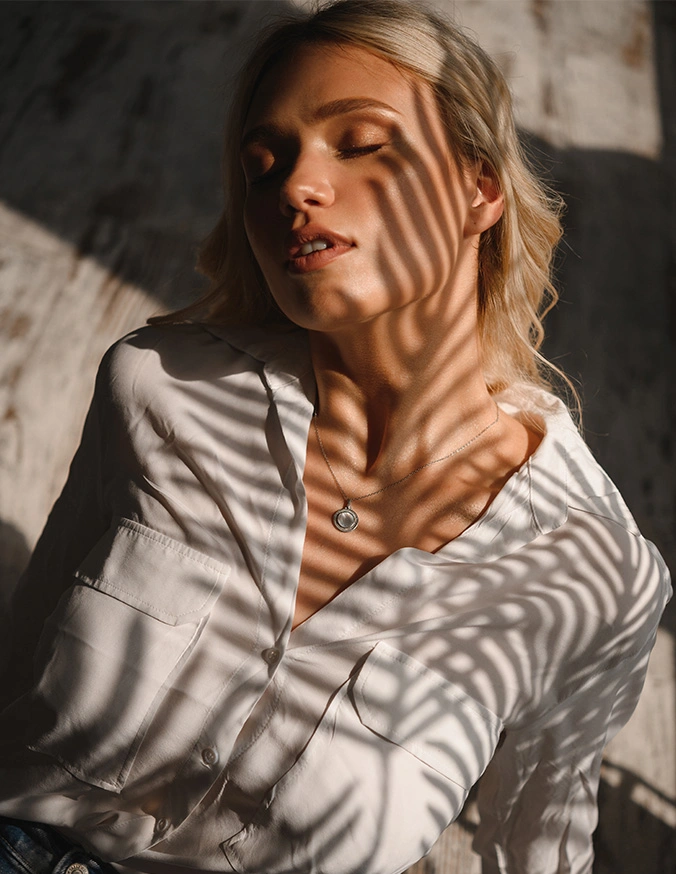 Woman in white shirt with shadow patterns from blinds across face and body in natural sunlight - Liposuction of the Neck and Chin in Las Vegas, NV
