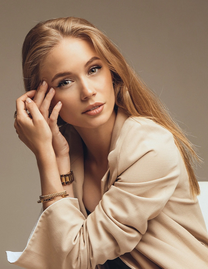 Elegant woman in beige blouse wearing gold jewelry posing with hand near face in professional studio portrait - Lower Eyelid Surgery with Fat Repositioning in Las Vegas, NV