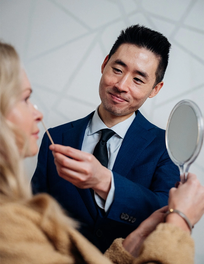 Harry Ching MD in suit consulting with blonde female patient while holding mirror in office - Rhinoplasty in Las Vegas, NV