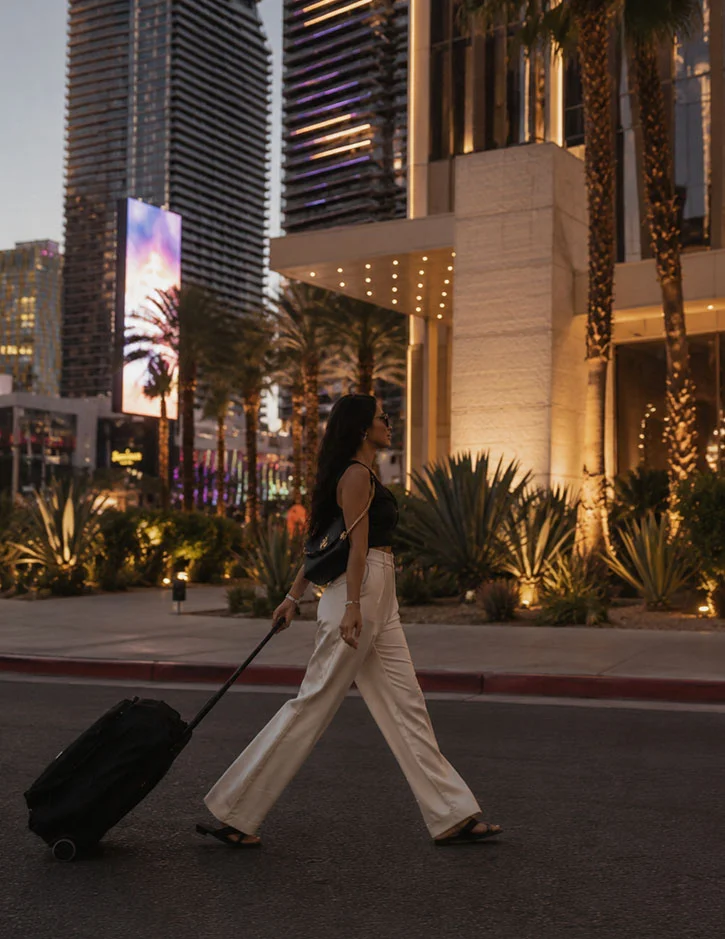 Stylishly dressed woman pulling a rolling suitcase along a palm-lined boulevard in Las Vegas at dusk. Modern luxury towers, illuminated landscaping, and glowing architectural lighting create an upscale urban resort atmosphere.