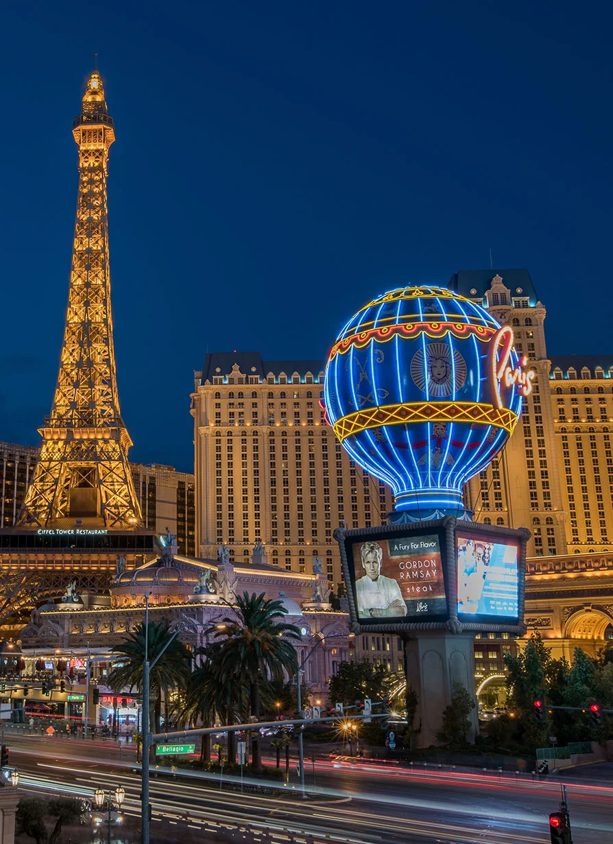 Nighttime view of the Las Vegas Strip featuring illuminated Paris Las Vegas landmarks, including the replica Eiffel Tower and brightly lit hot air balloon sign. Neon lights, luxury hotels, and streaking traffic create a vibrant cityscape against the dark evening sky.